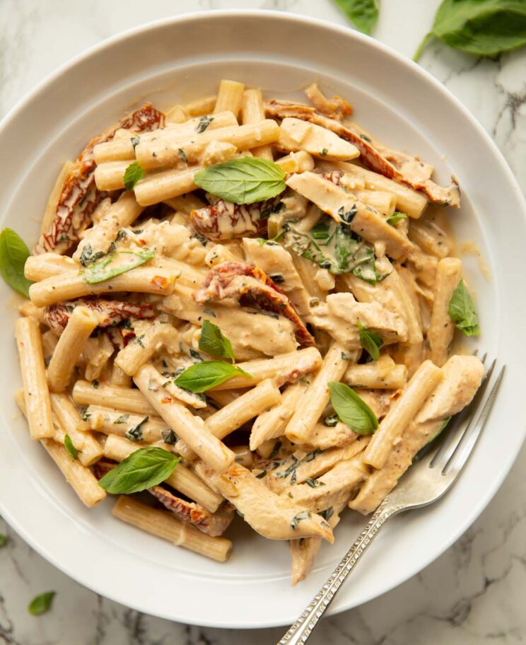 overhead shot of chicken sun dried tomato pasta in white dish with silver fork garnished with fresh basil