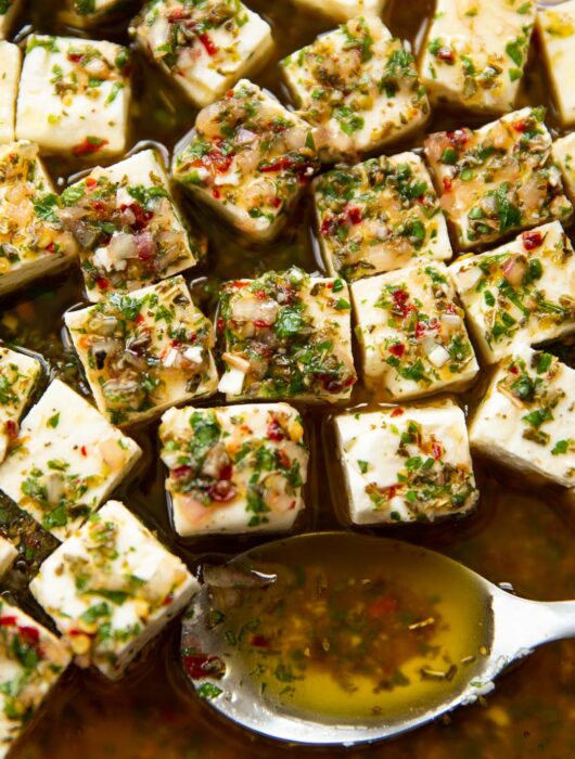 close up shot of marinated feta in glass dish with silver spoon