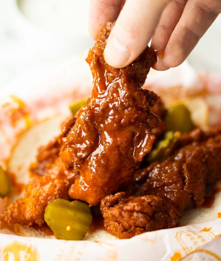hand lifting chicken tender from basket with 2 other tenders and pickles