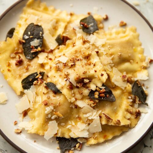 close up shot of ravioli on small white plate garnished with crispy sage, pecans and parmesan