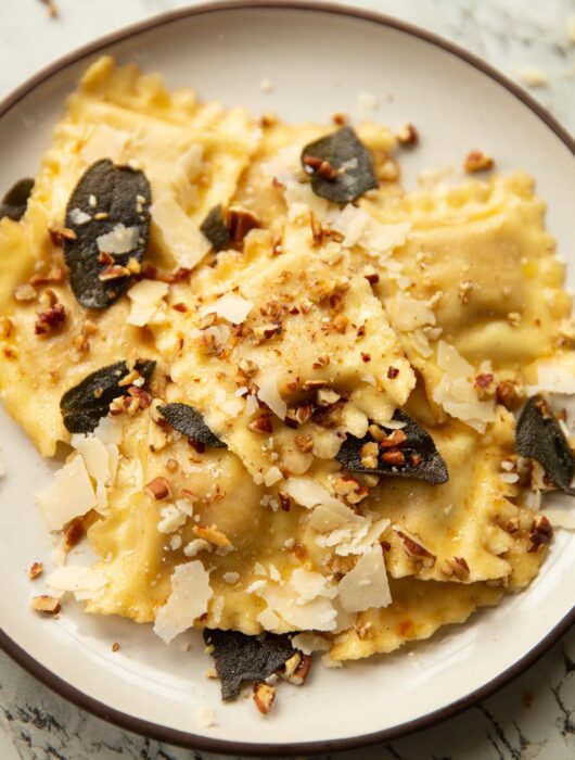 close up shot of ravioli on small white plate garnished with crispy sage, pecans and parmesan