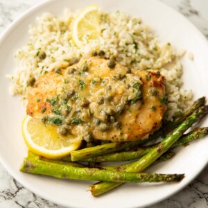 chicken piccata served on small white plate with rice, asparagus and lemon wedges