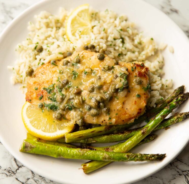 chicken piccata served on small white plate with rice, asparagus and lemon wedges