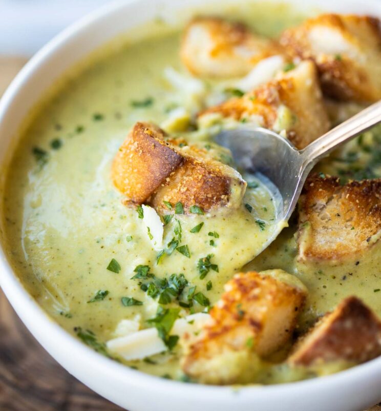 close up shot of silver spoon digging into small white bowl of broccoli soup