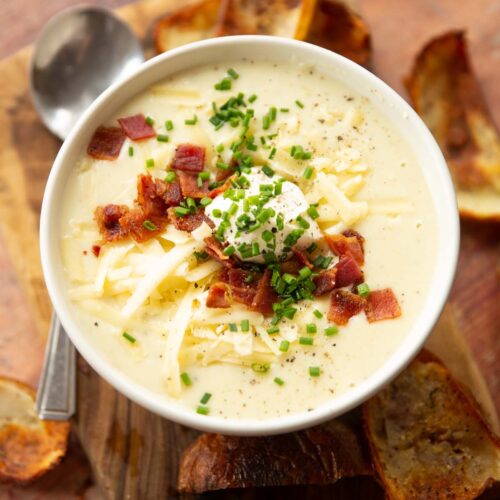 loaded baked potato soup in small white bowl on wooden chopping board surrounded by crispy potato skins