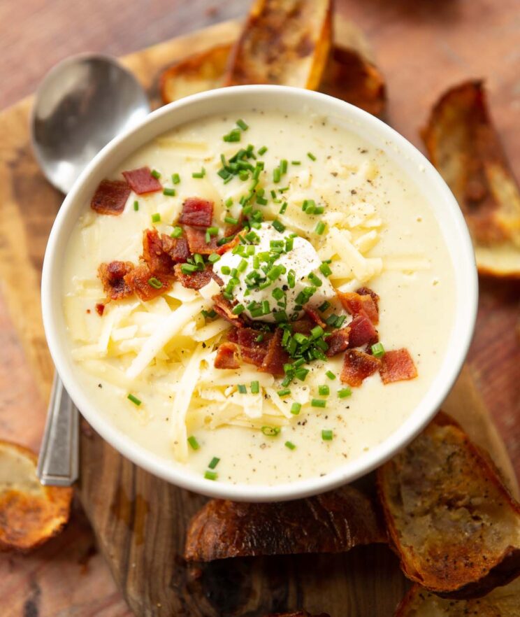 loaded baked potato soup in small white bowl on wooden chopping board surrounded by crispy potato skins