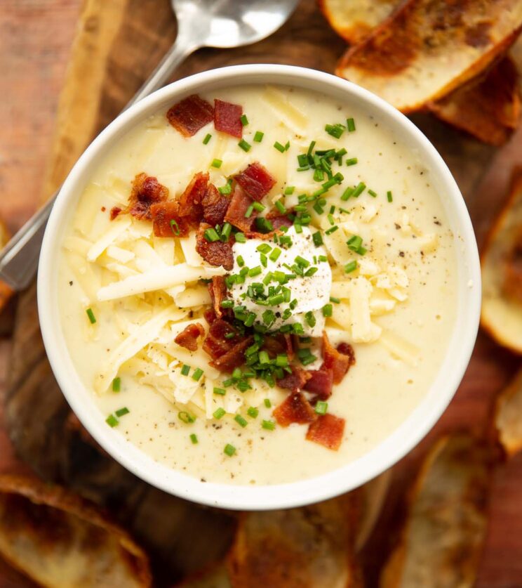 close up overhead shot of loaded potato soup in small white bowl on wooden backdrop surrounded by crispy potato skins
