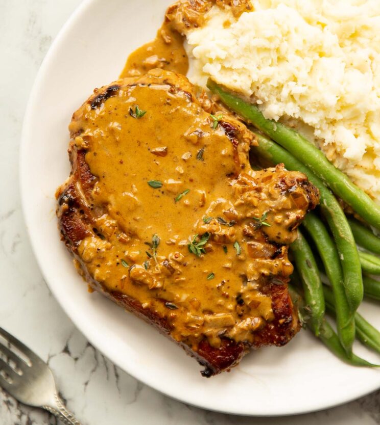 close up overhead shot of pork chop with garlic sauce on white plate with green beans and mashed potatoes