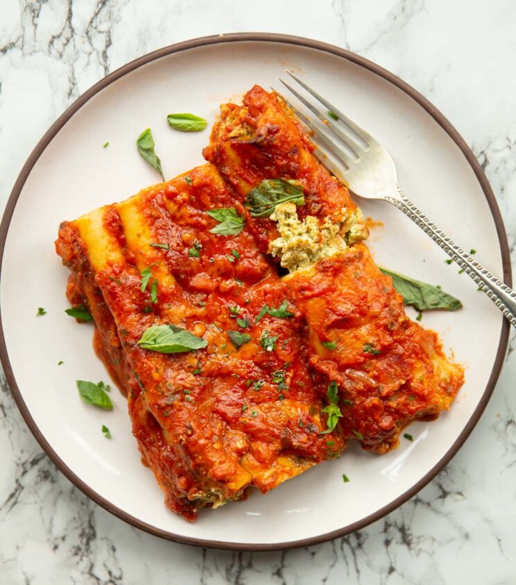 overhead shot of 3 ricotta pesto cannelloni on small white plate with silver fork on marble background