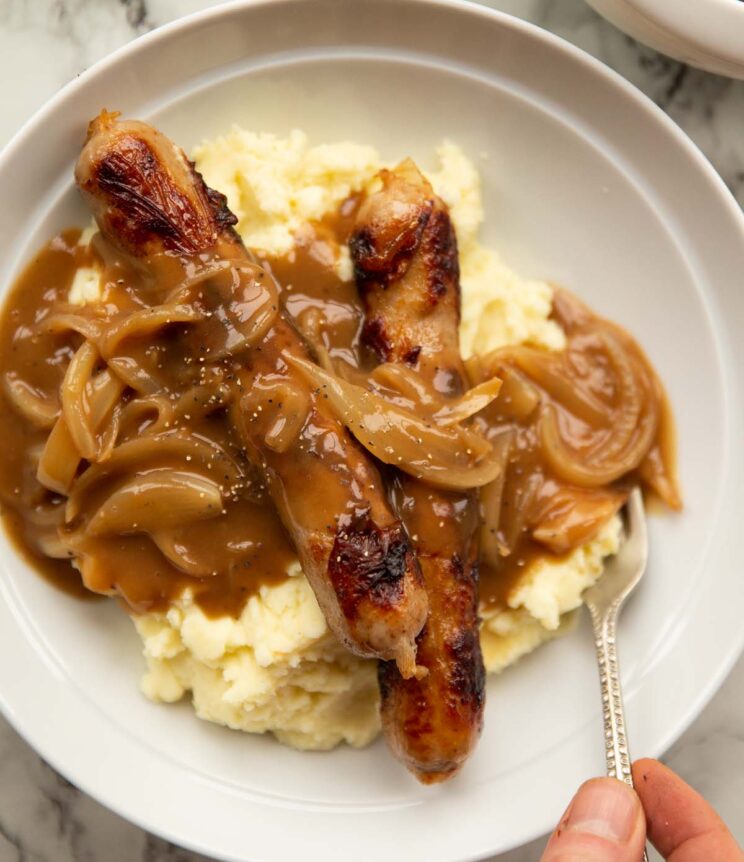 overhead shot of bangers and mash in large white bowl with onion gravy and hand digging in with silver fork