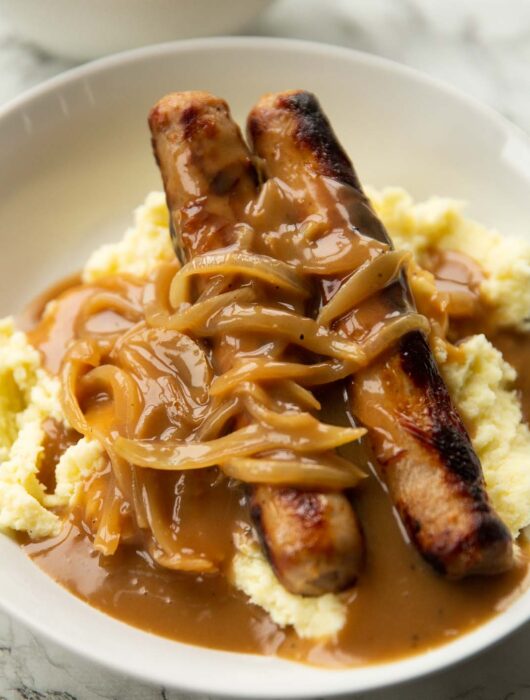 2 sausages on a bed of mashed potatoes with onion gravy in large white bowl
