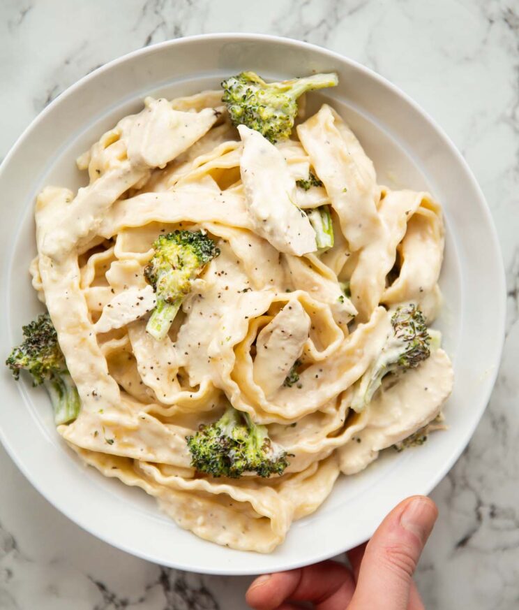 overhead shot of hand holding white dish with chicken and broccoli pasta in it
