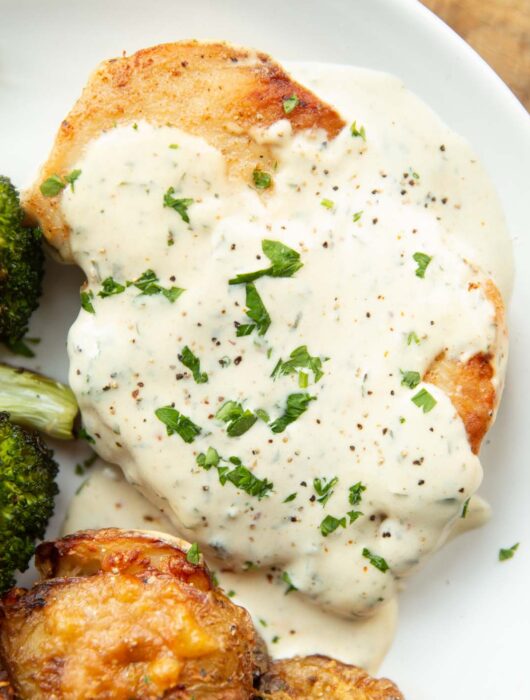 close up overhead shot of chicken breast and creamy garlic sauce on white plate with broccoli and smashed potatoes