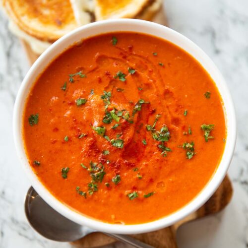 roasted red pepper soup in small white bowl on wooden chopping board with grilled cheese in background