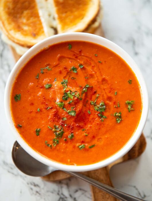 roasted red pepper soup in small white bowl on wooden chopping board with grilled cheese in background