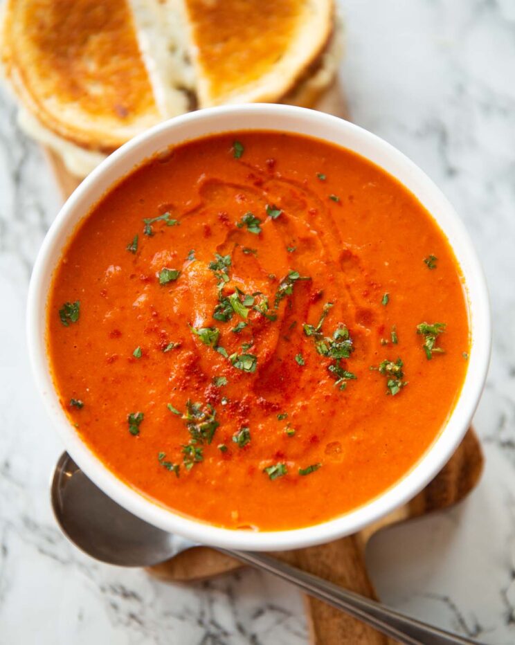 roasted red pepper soup in small white bowl on wooden chopping board with grilled cheese in background