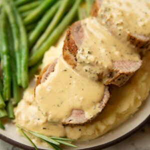 close up shot of pork tenderloin slices on mashed potato with mustard sauce and green beans blurred in background