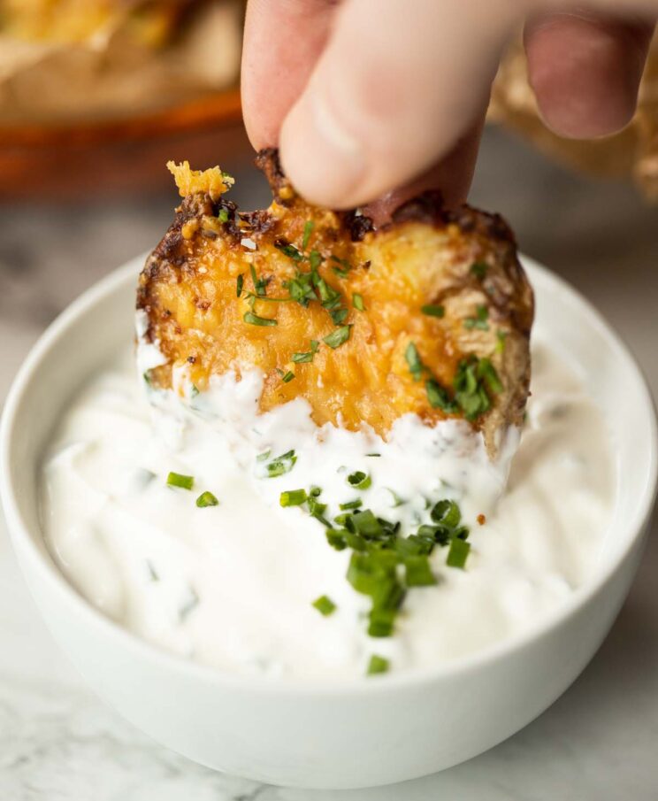 close up shot of hand dunking smashed potatoes parmesan into small white pot of dip