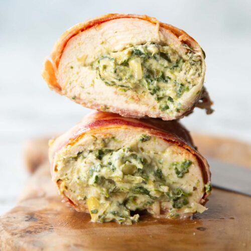 close up side shot showing spinach and artichoke filling inside chicken breasts wrapped in bacon on chopping board