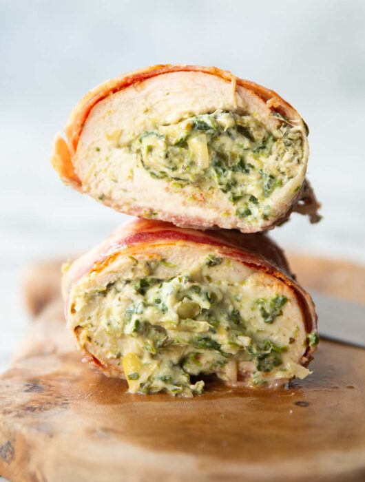 close up side shot showing spinach and artichoke filling inside chicken breasts wrapped in bacon on chopping board