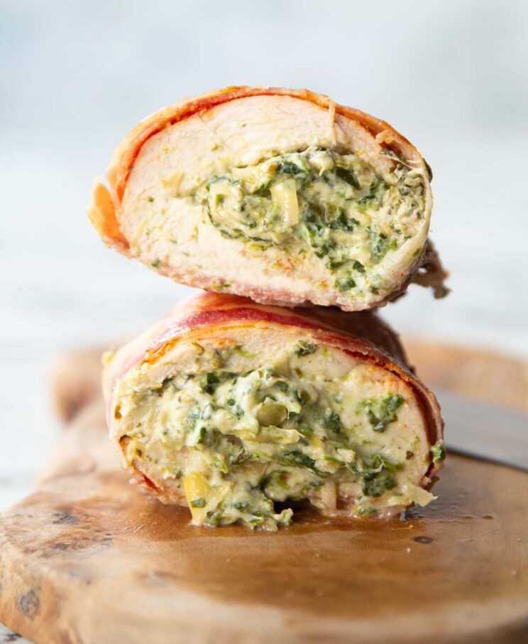 close up side shot showing spinach and artichoke filling inside chicken breasts wrapped in bacon on chopping board