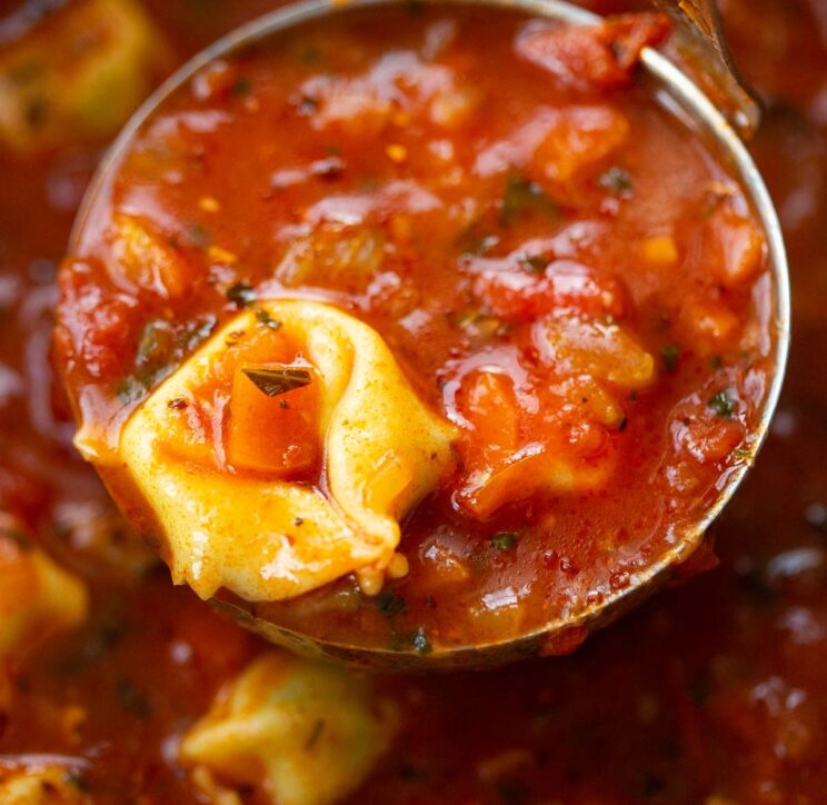 close up shot of tomato and tortellini soup in silver ladle