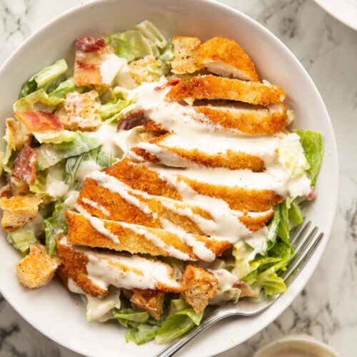 overhead shot of chicken caesar salad in large white bowl on marble backdrop with silver fork