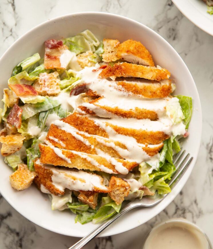 overhead shot of chicken caesar salad in large white bowl on marble backdrop with silver fork