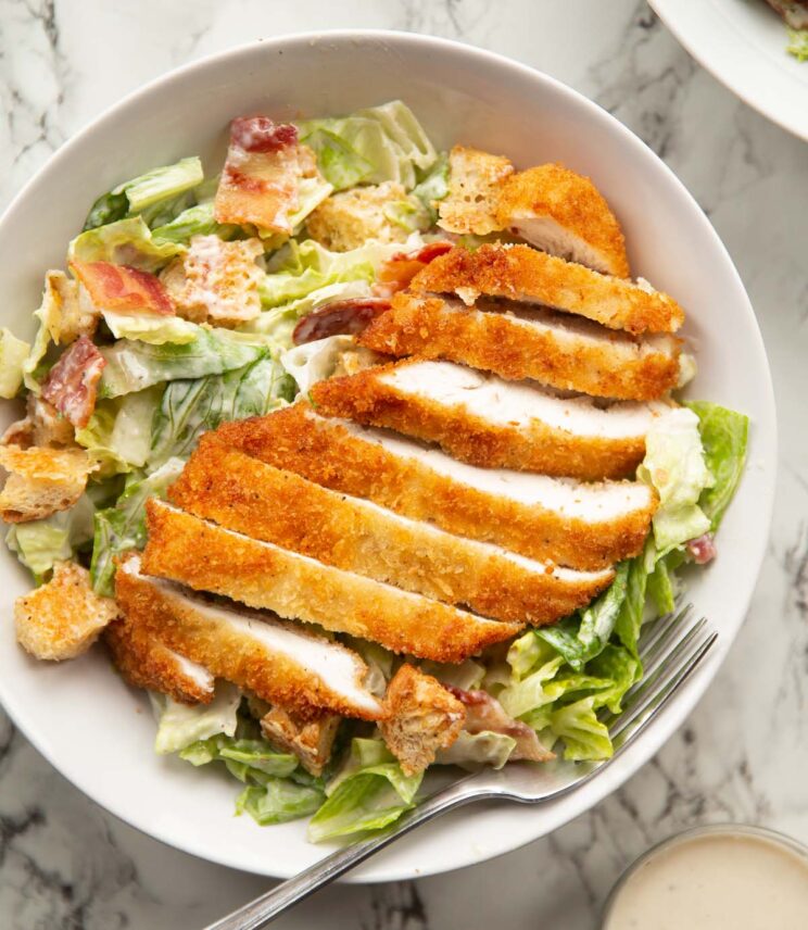overhead shot of crispy chicken caesar salad in large white dish with silver fork on marble background