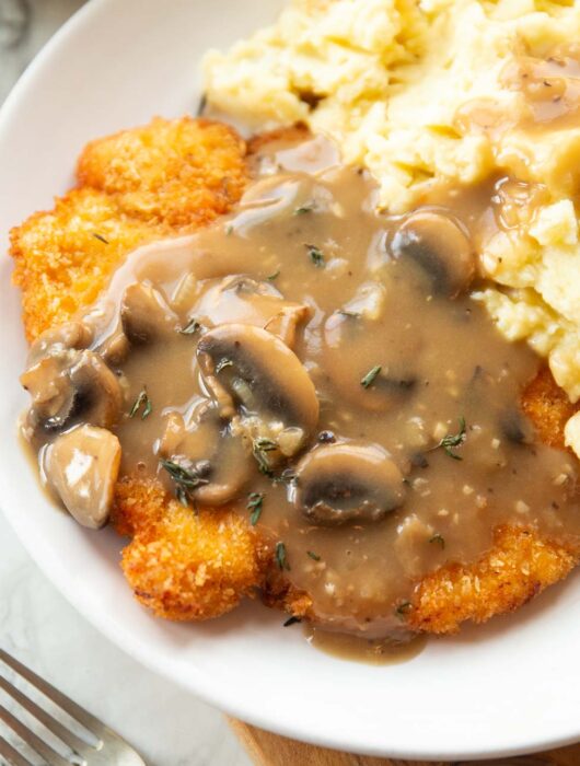 close up shot of chicken schnitzel with mushroom gravy and mashed potatoes on white plate