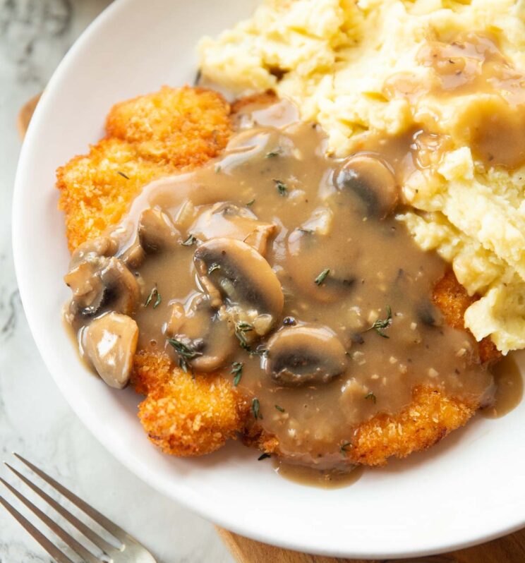 close up shot of chicken schnitzel with mushroom gravy and mashed potatoes on white plate