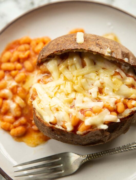 overhead shot of jacket potato with baked beans and melted cheddar cheese on top on small white plate with silver fork