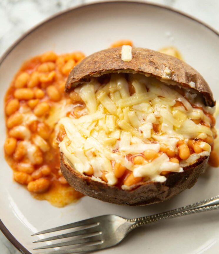 overhead shot of jacket potato with baked beans and melted cheddar cheese on top on small white plate with silver fork