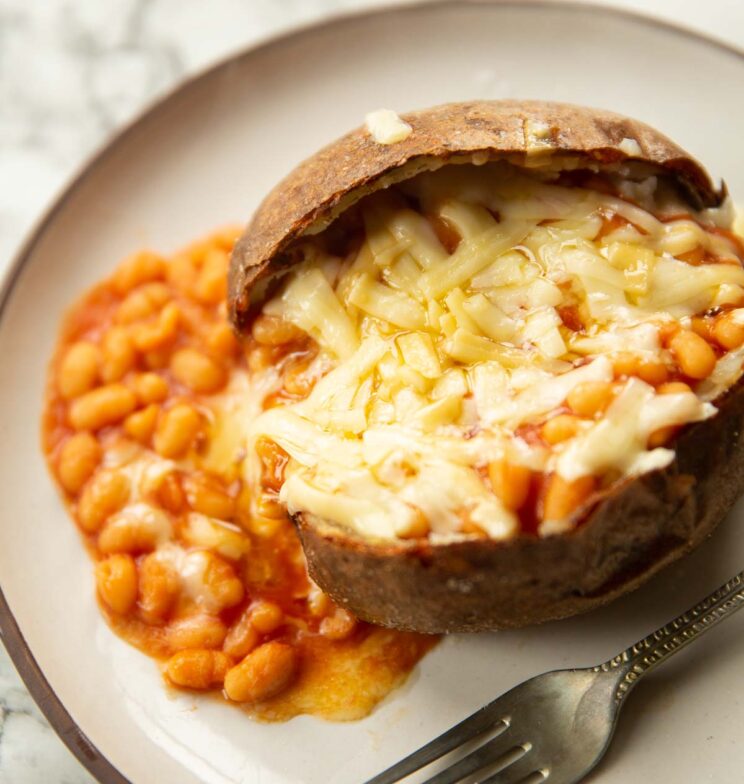 close up shot of jacket potato with beans and cheese on small white plate with silver fork to one side