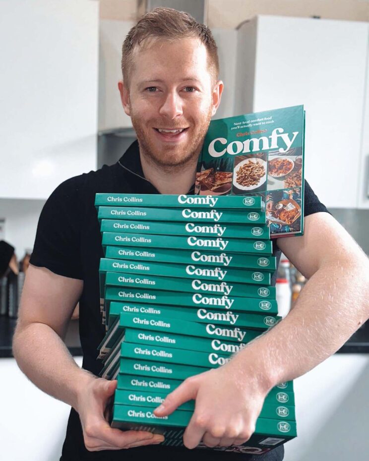 Chris Collins holding stack of Comfy cookbook in kitchen