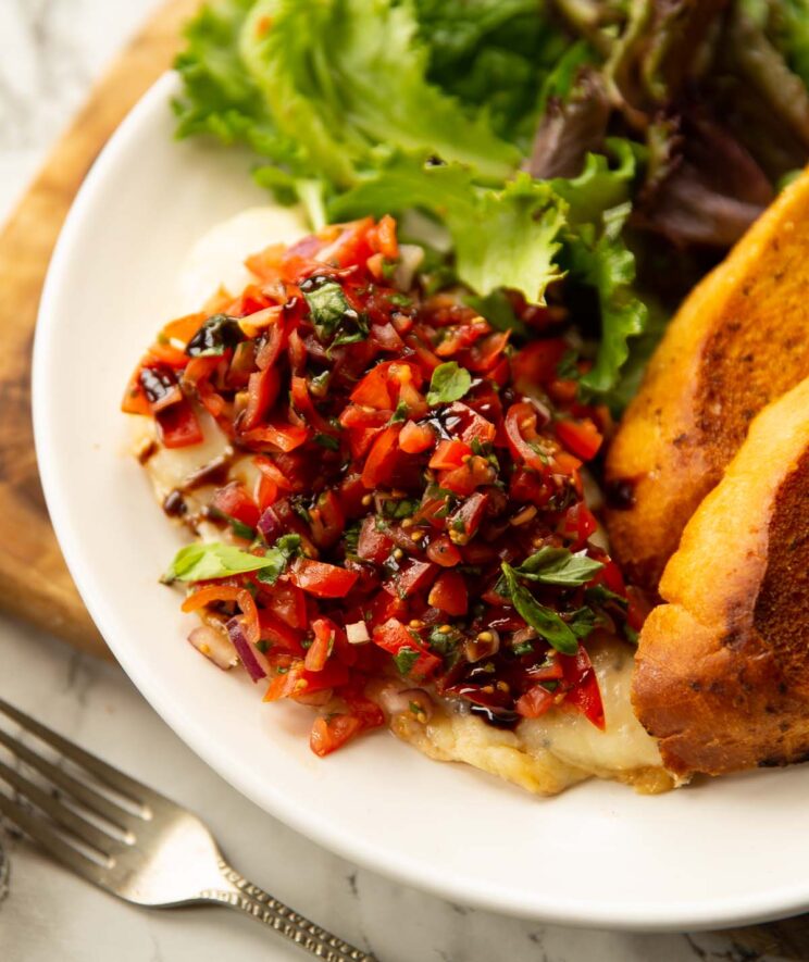 cheesy bruschetta chicken on a small white plate with salad and garlic bread