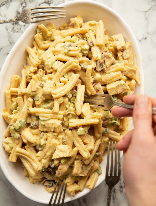 overhead shot of coronation chicken and bacon pasta salad in large oval dish with forks surrounding