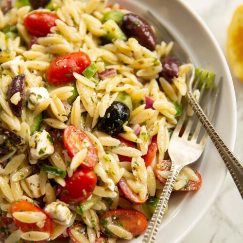 close up shot of greek orzo pasta salad in white bowl with 2 silver forks