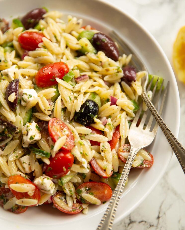 close up shot of greek orzo pasta salad in white bowl with 2 silver forks
