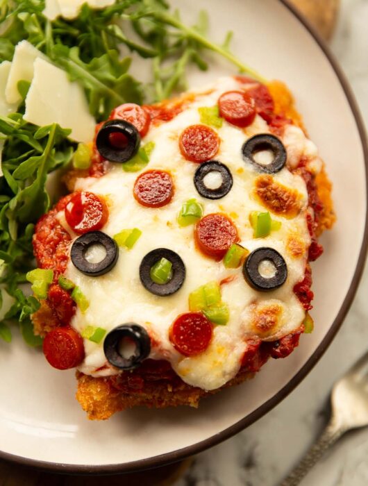 close up shot of pizza chicken parmesan on small white plate with rocket salad