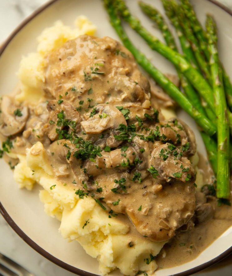 close up shot of chicken marsala on mashed potato with asparagus on small white plate