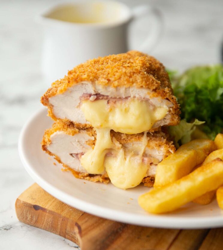 two baked chicken cordon bleu halves stacked on each other on small white plate with chips and salad