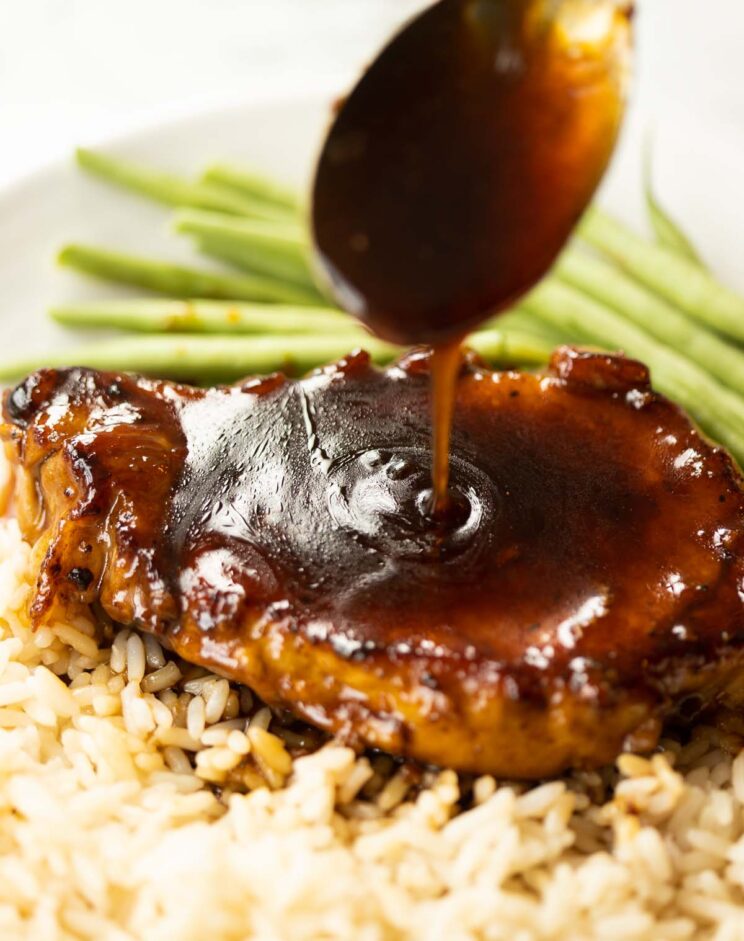 spoon drizzling brown sugar glaze over pork chop on white plate with rice and green beans