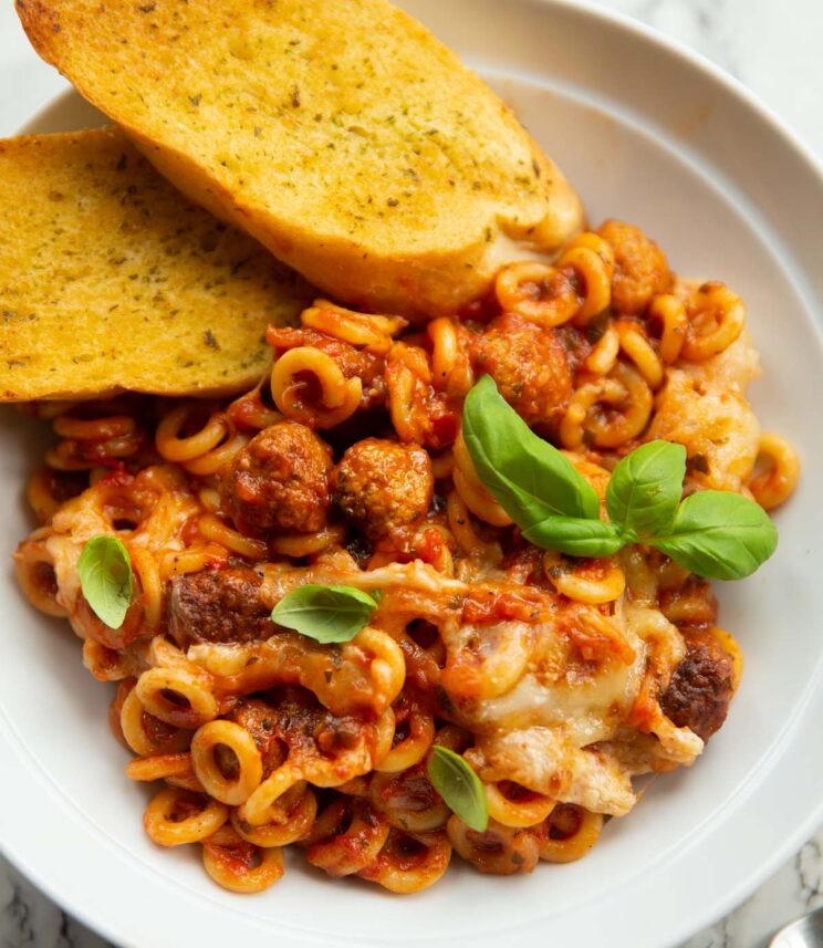 cheesy sausage spaghetti hoops in large white bowl with garlic bread garnished with basil