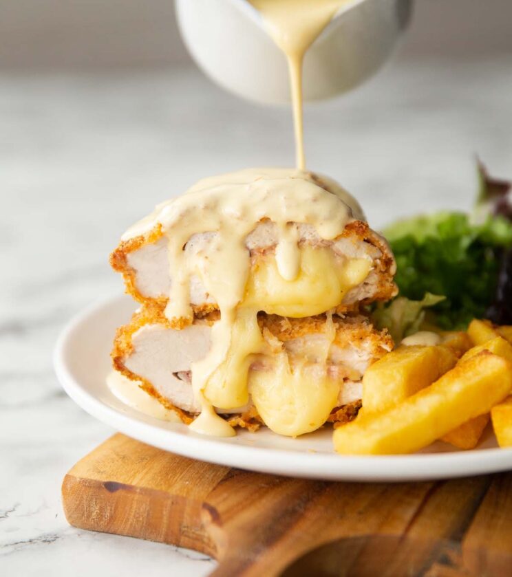 jug pouring sauce over chicken cordon bleu on small white plate with chips and salad