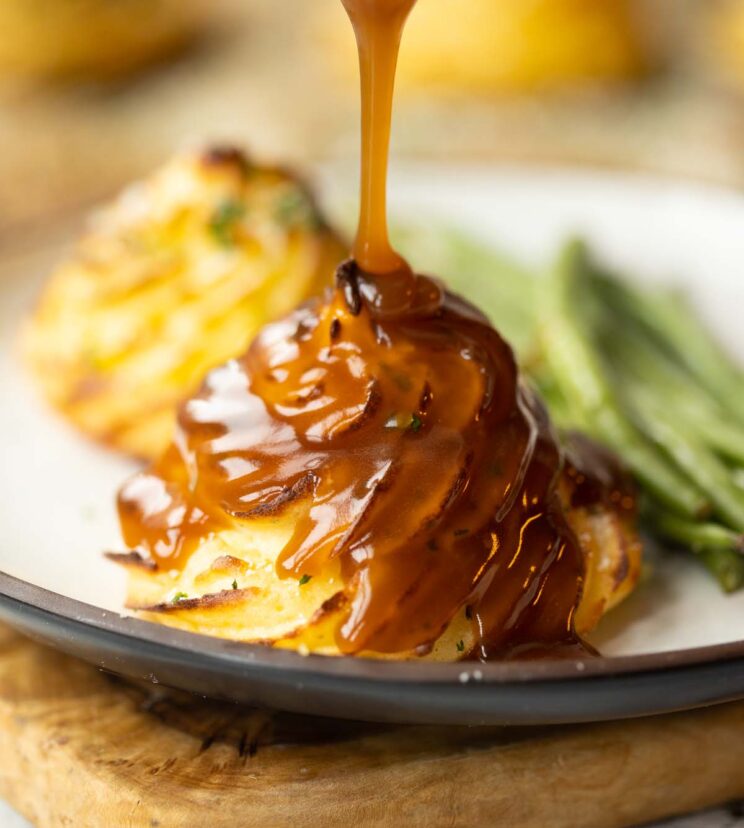 pouring gravy over a duchess potato on small white plate with green beans