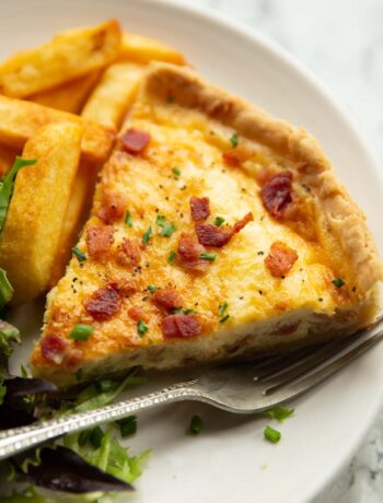 close up shot of slice of quiche lorraine on small white plate with chips and salad