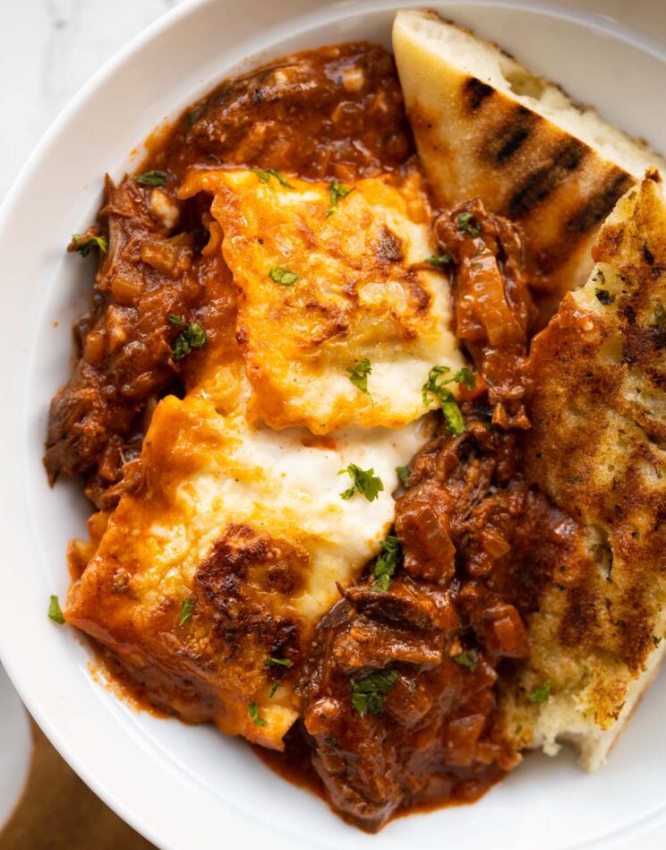 close up overhead shot of beef ragu ravioli bake in large white bowl with garlic bread