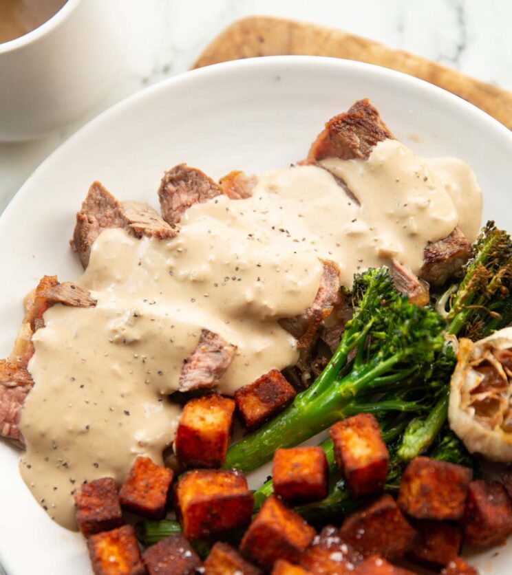 close up shot of sliced steak coated in sauce on white plate with broccolini and potatoes