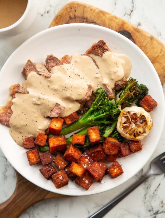 sliced steak with creamy garlic sauce on white plate with potatoes and broccolini